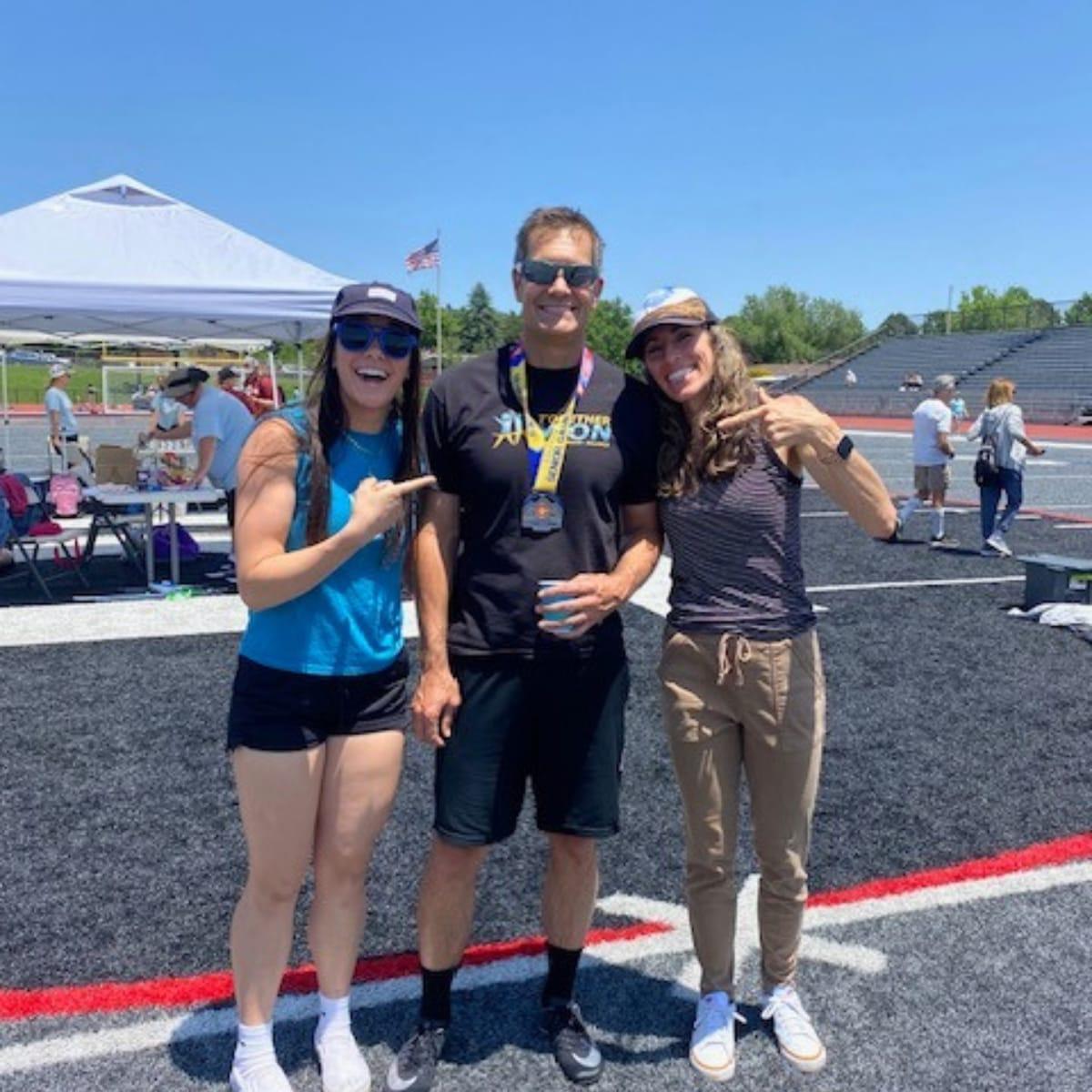Aaron wearing his Senior Games medal, flanked by his two daughters pointing at him on the track infield