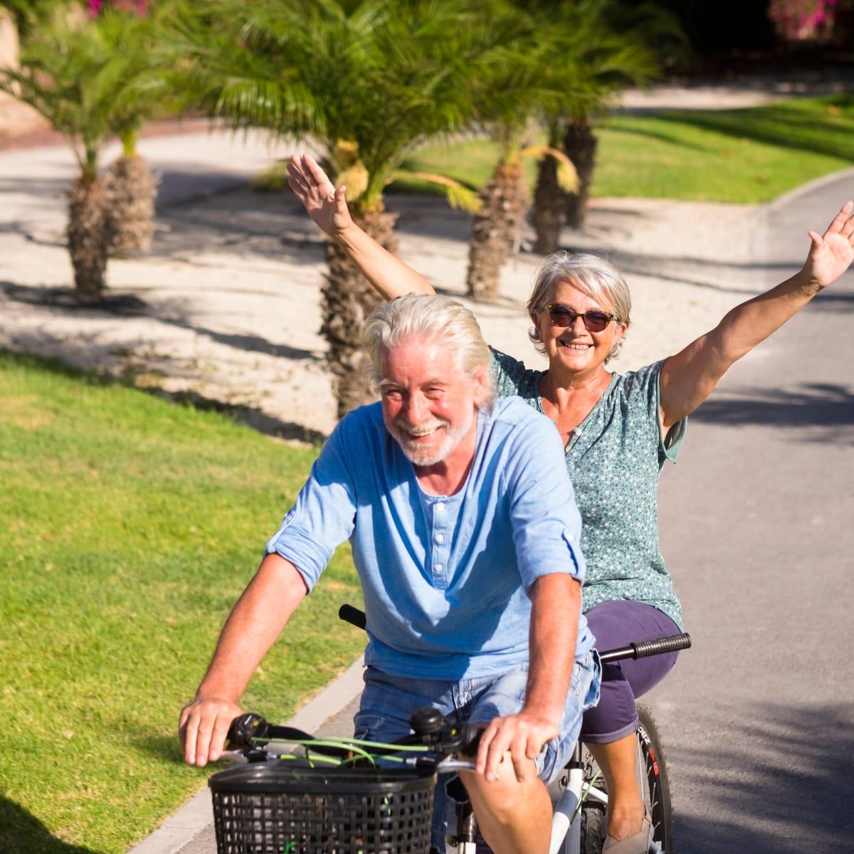 A smiling older couple riding a tandem bicycle past palm trees, the woman waving her arms