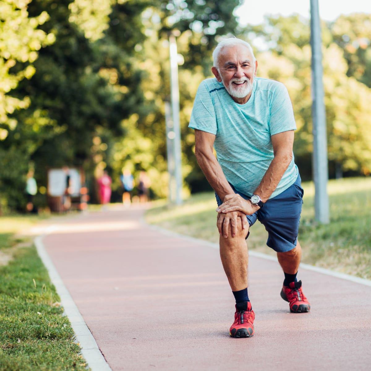 A smiling older man in a teal T-shirt doing a forward lunge stretch on a park path