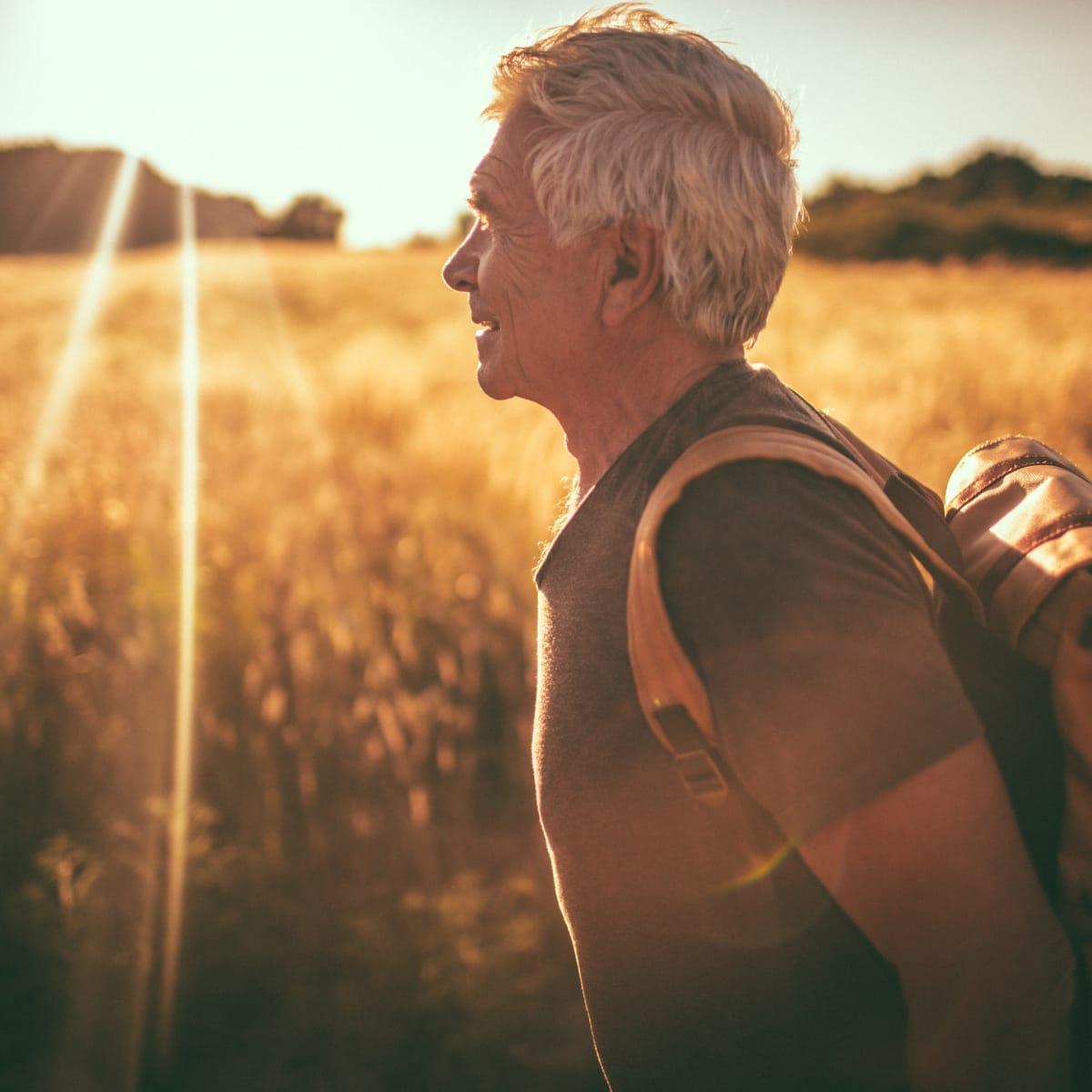 An older man with a backpack walking through a golden field at sunset