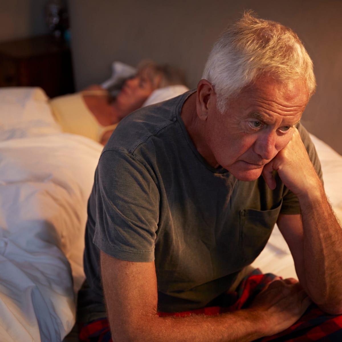 An older man sitting on the edge of the bed at night, looking worried, while his partner sleeps