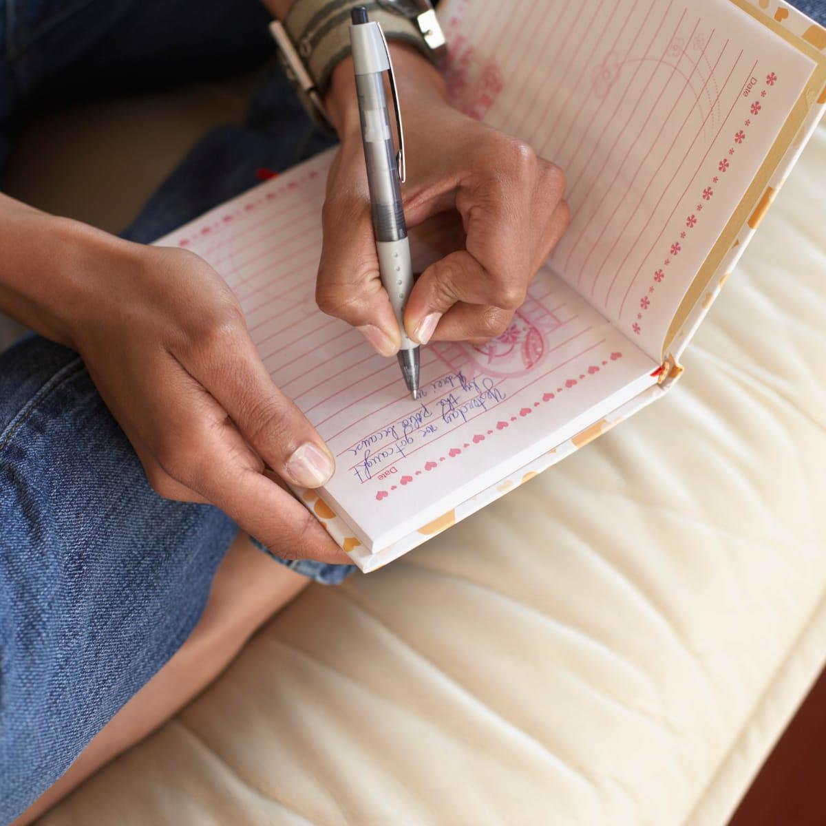 A person writing in a small notebook with a pen while sitting on a bed