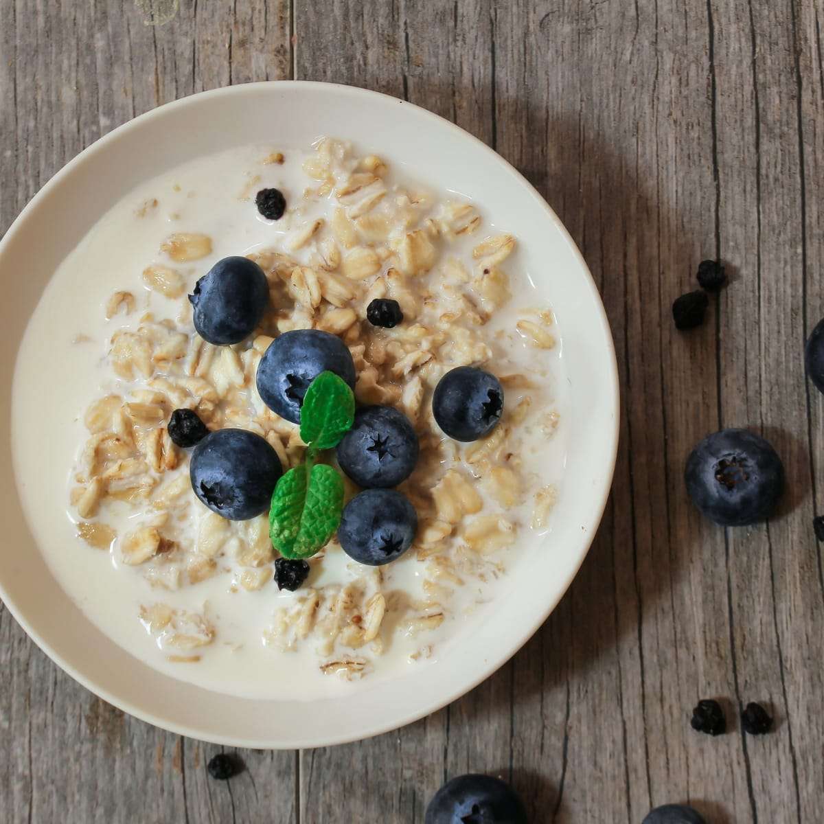 A white bowl of oats and milk topped with fresh blueberries and a mint leaf on a wooden table