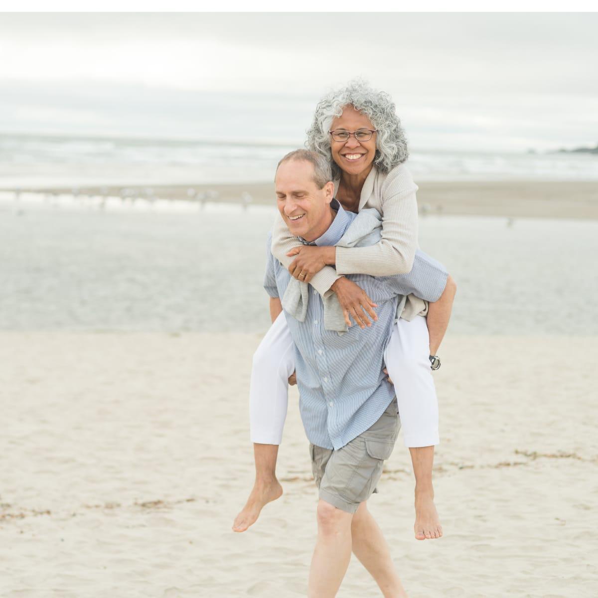 An older man laughing as he gives his wife a piggyback ride along a sandy beach