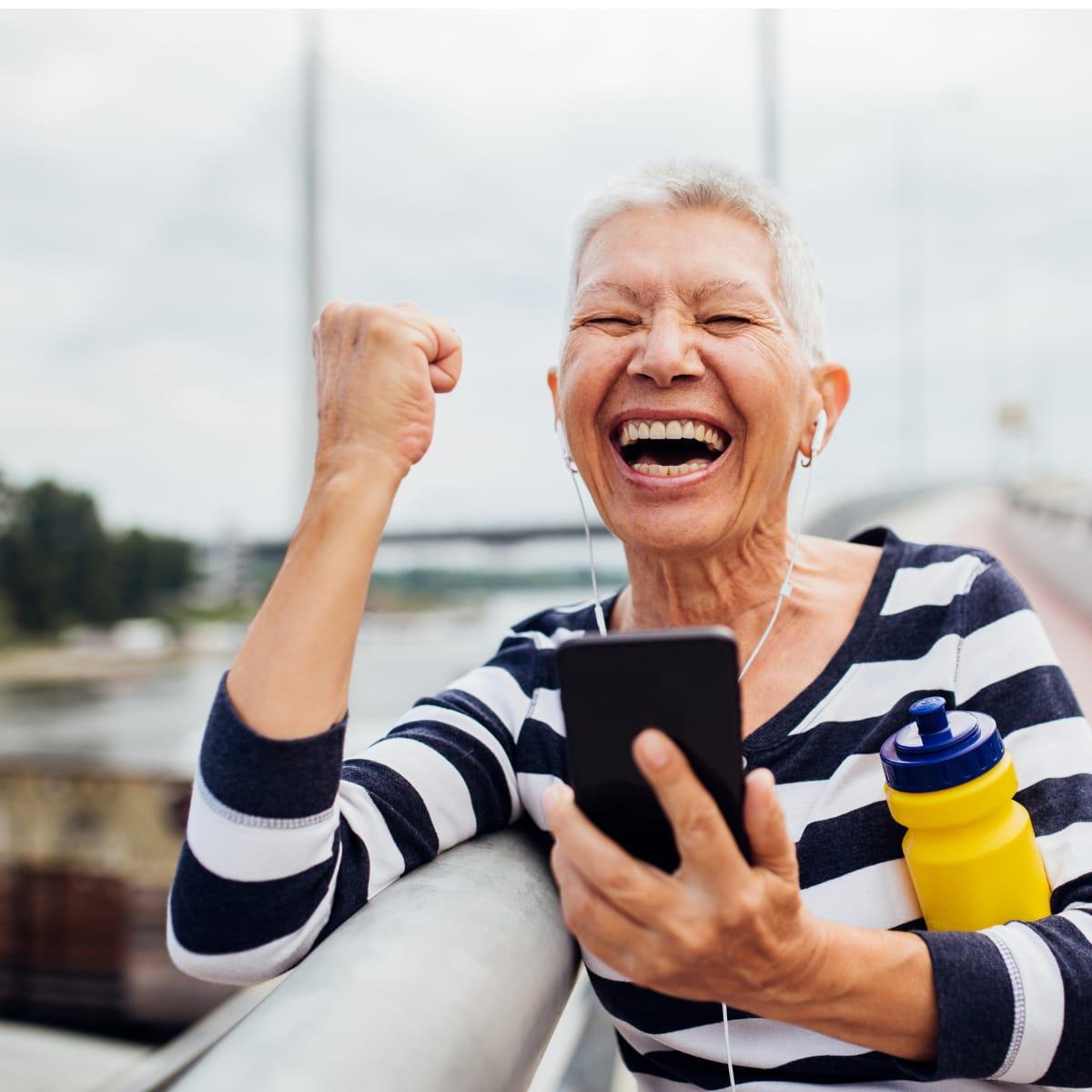 An older woman in earbuds cheering with a raised fist while checking her phone on a bridge
