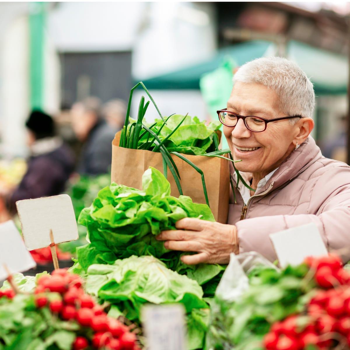 A smiling older woman choosing fresh lettuce at an outdoor market, a paper grocery bag on her arm