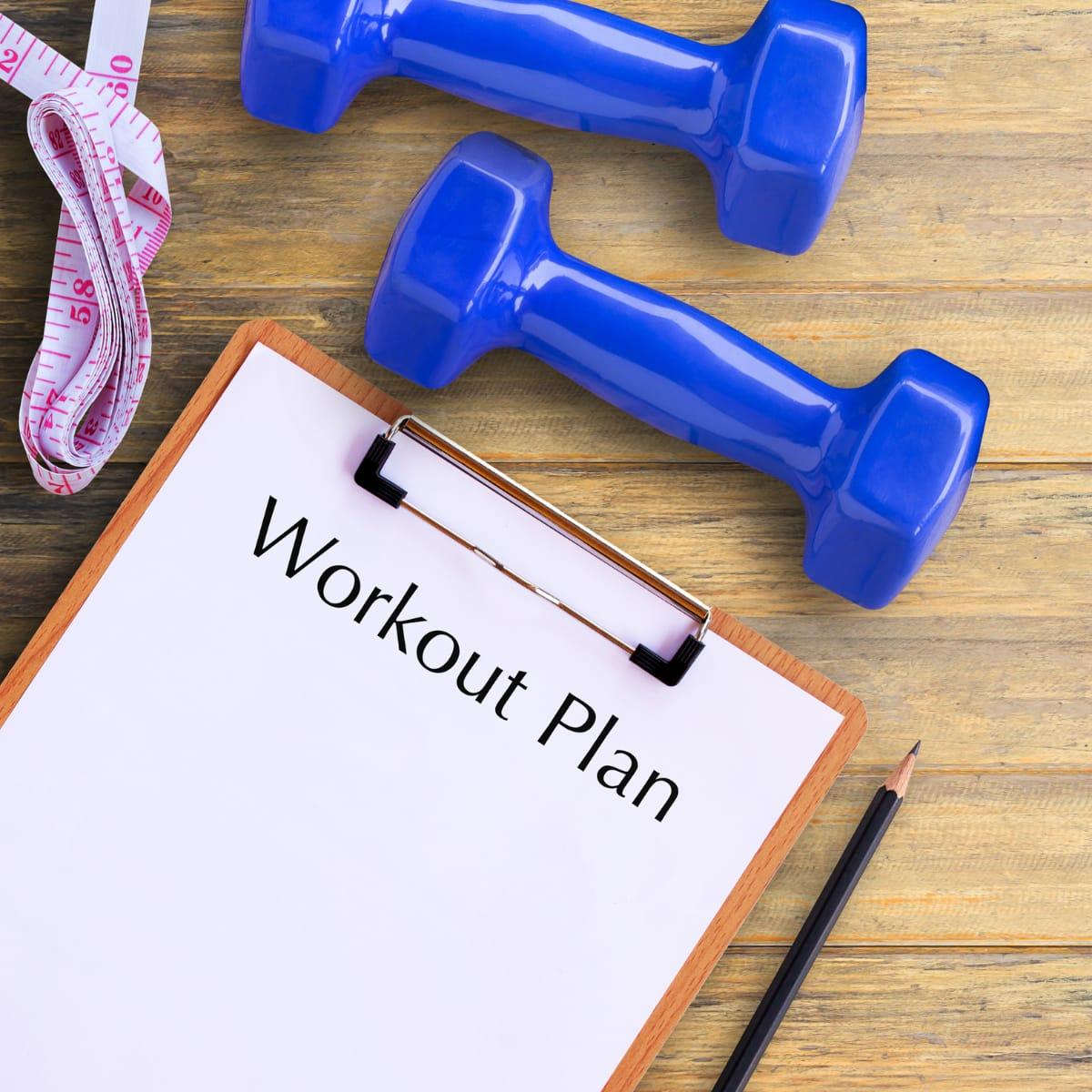 A clipboard labeled "Workout Plan" next to two blue dumbbells, a pencil and a tape measure on a wooden table
