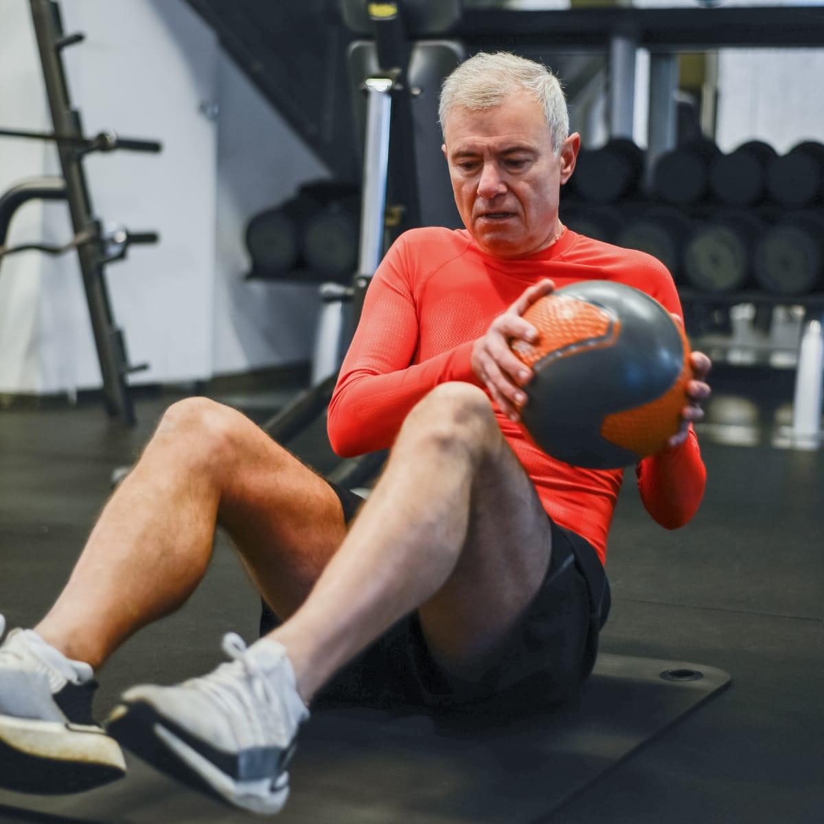 An older man in a red athletic shirt doing a seated twist with a medicine ball on a gym floor