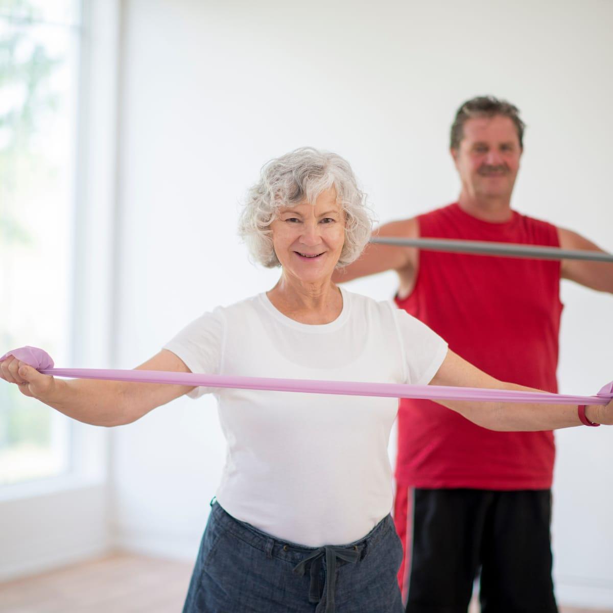 An older woman stretching a resistance band in front of her chest while a man works with a bar behind her