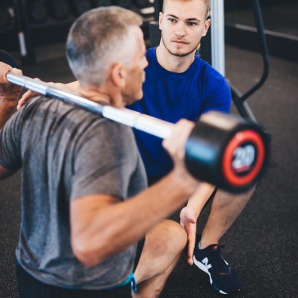 A trainer kneeling beside an older man and watching his form as he squats with a barbell on his shoulders