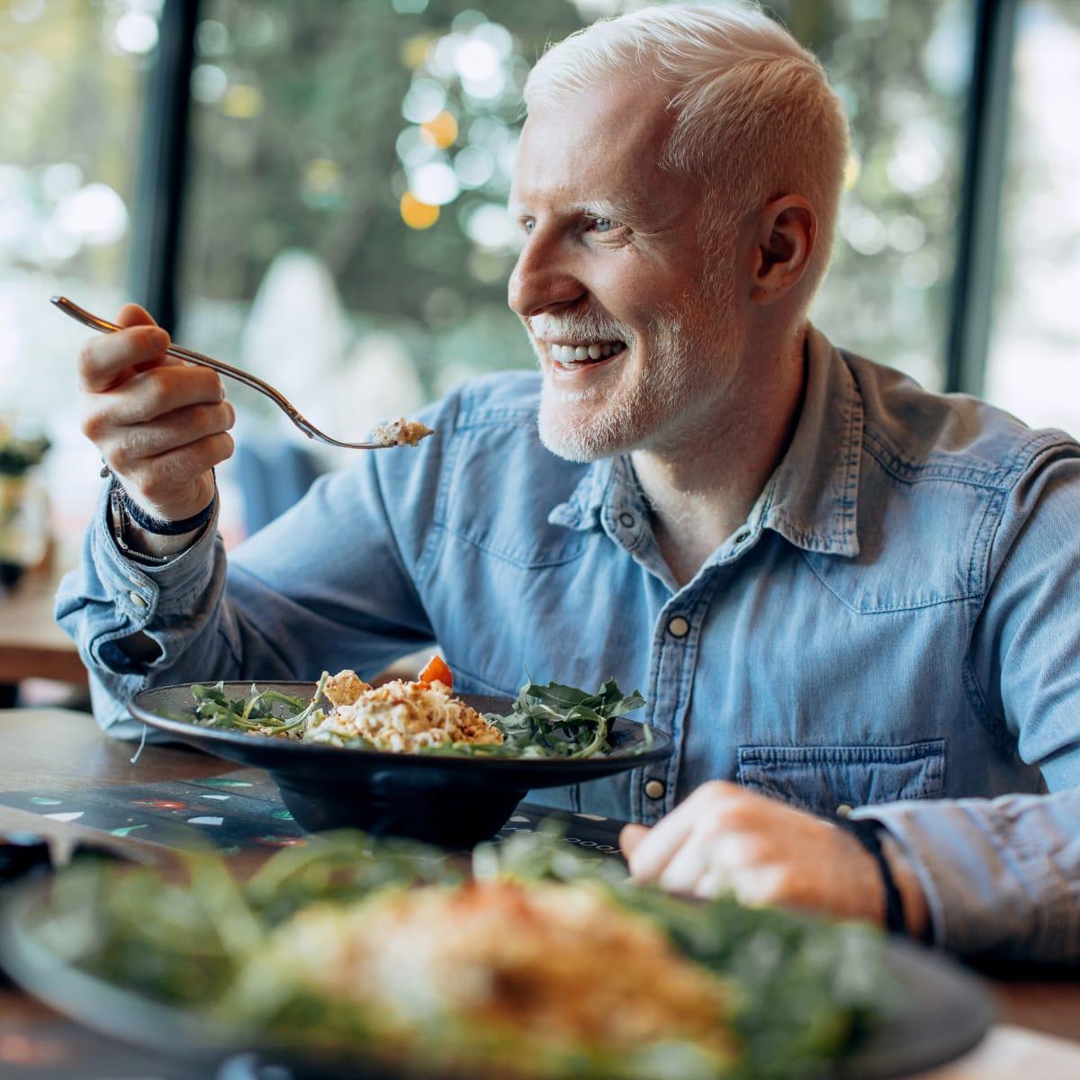 A smiling older man with a white beard eating a plate of salad at a restaurant table