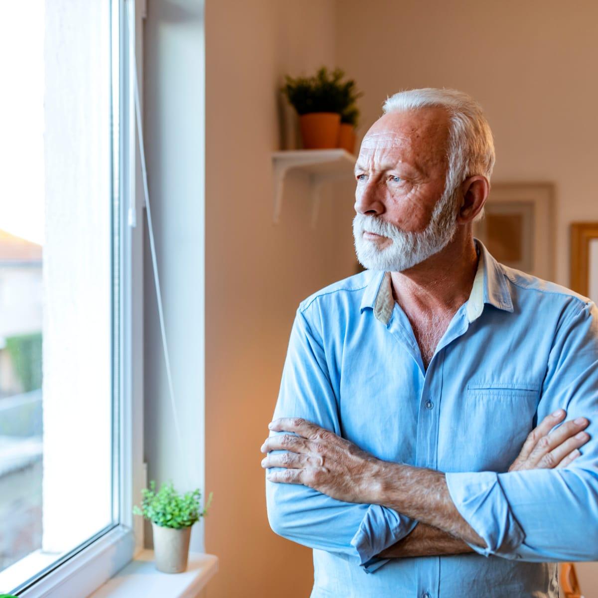 A thoughtful older man with a white beard standing by a window with his arms crossed