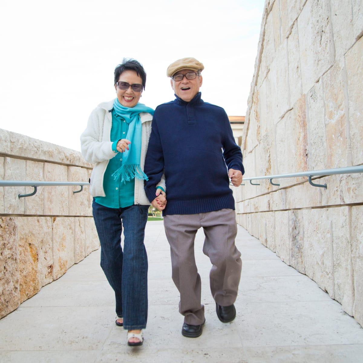 An older couple in a flat cap and teal scarf walking briskly arm in arm along a stone walkway