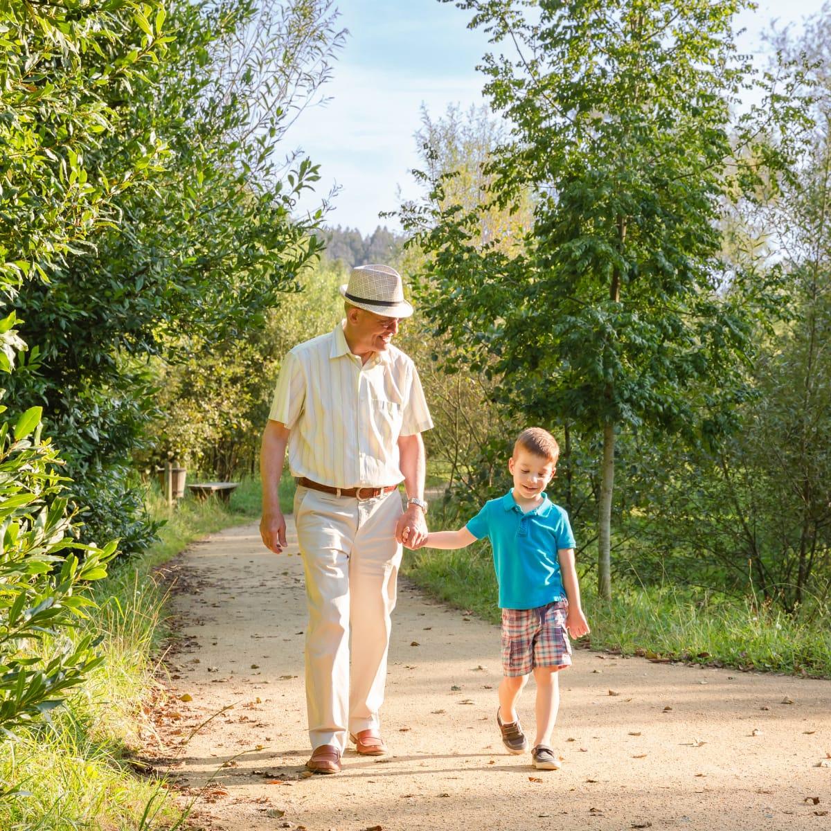 An older man in a sun hat walking hand in hand with his young grandson on a tree-lined dirt path
