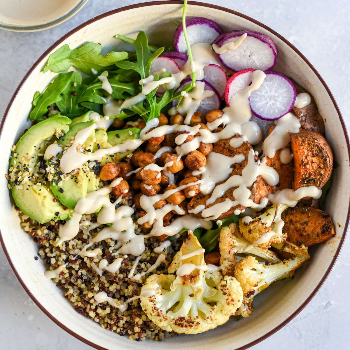 A quinoa bowl with roasted sweet potato, chickpeas, cauliflower, greens, avocado and radishes under a creamy drizzle