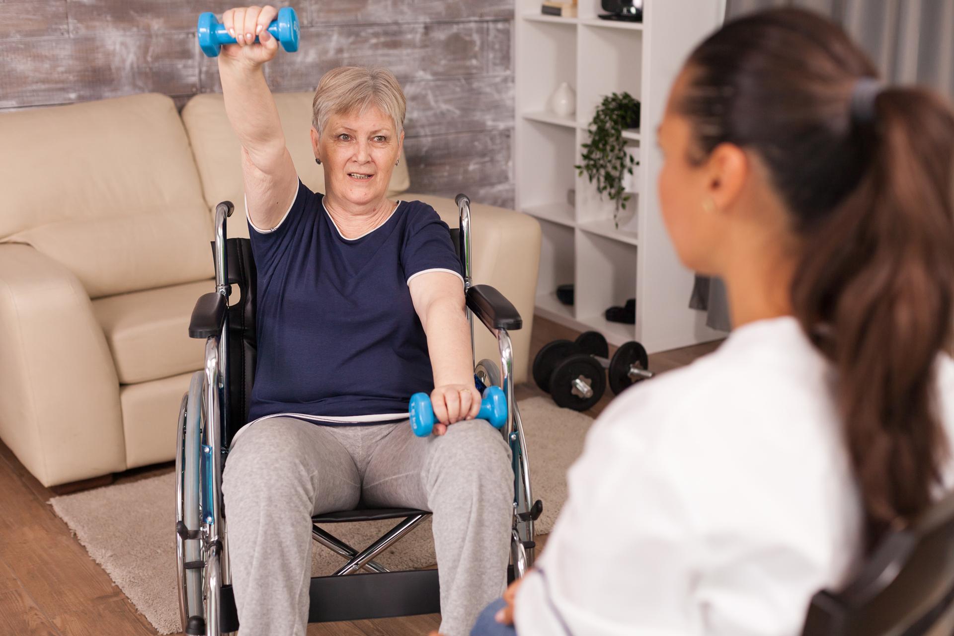 An older woman seated in a wheelchair raising her arms for shoulder exercises as a trainer coaches her at home
